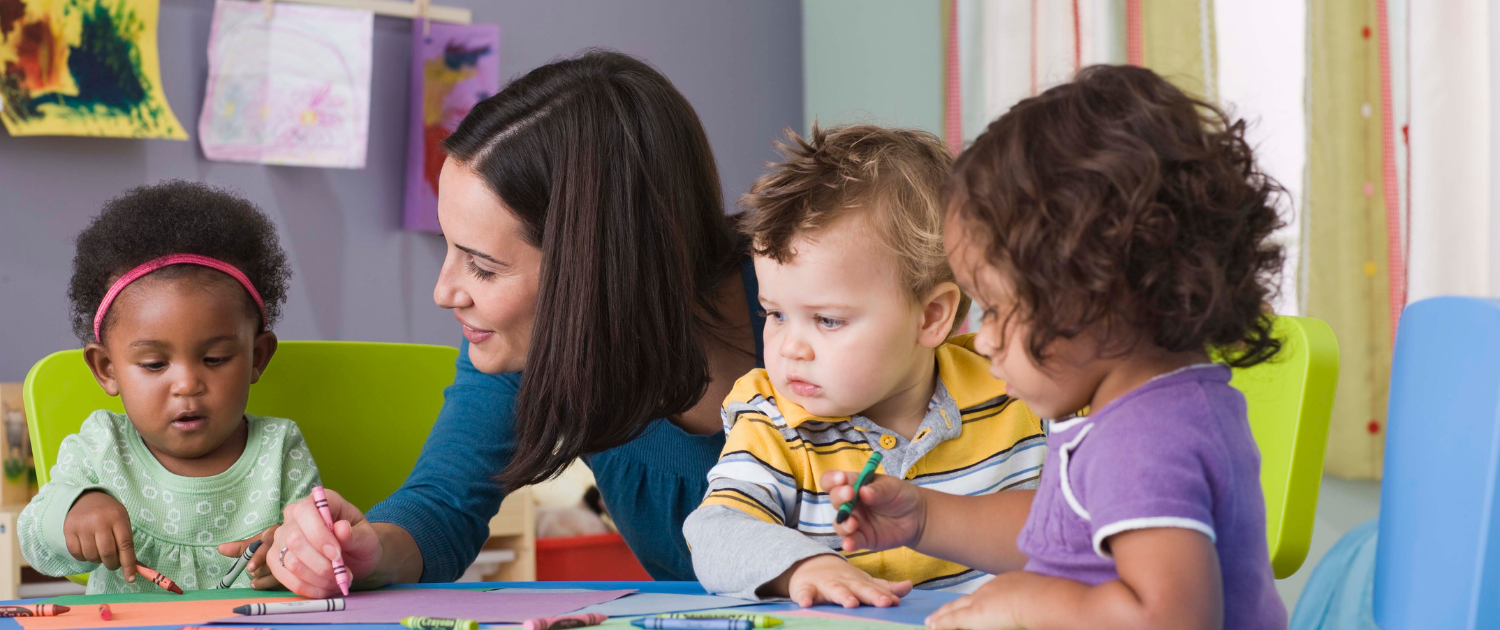 family child care provider sitting and smiling with small children at table