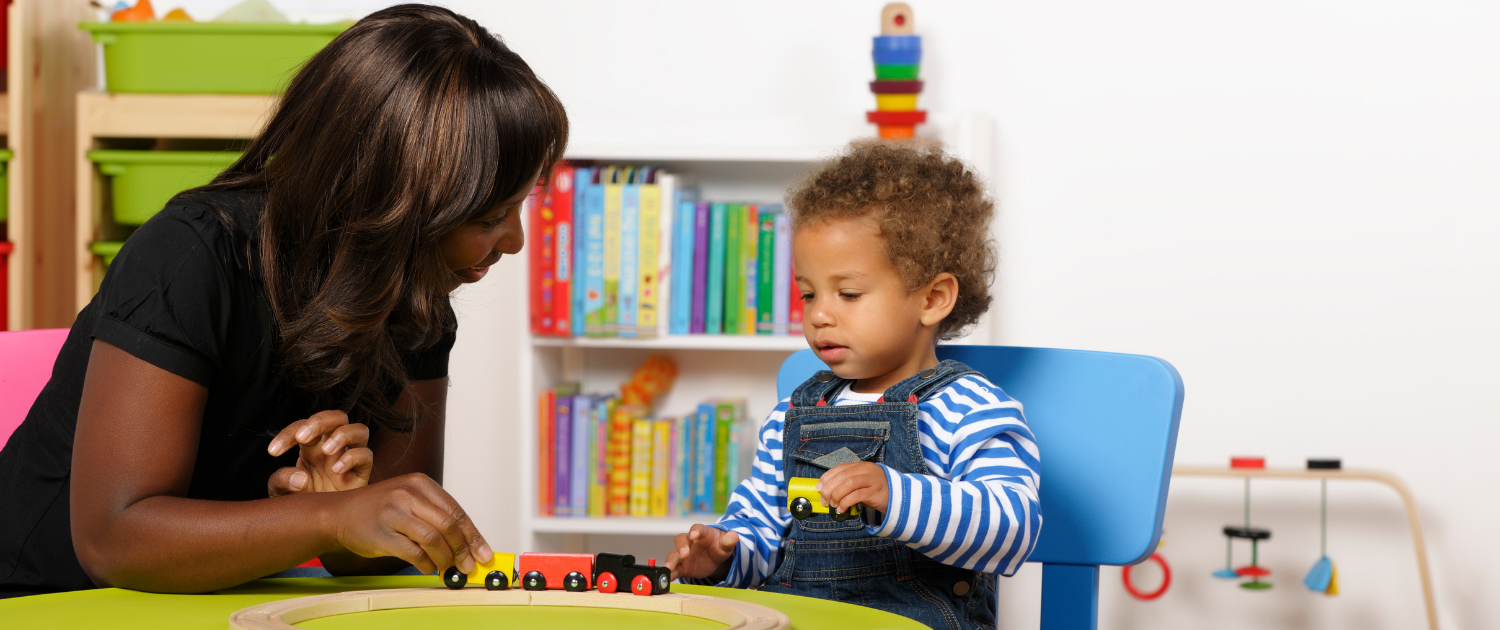 Teacher plays with a toy wooden train at a desk with a young child