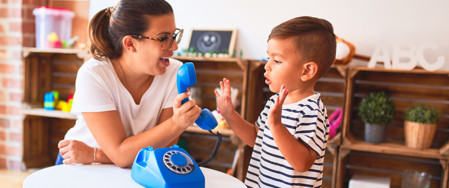 Teacher holds up the receiver of a play phone to a young child