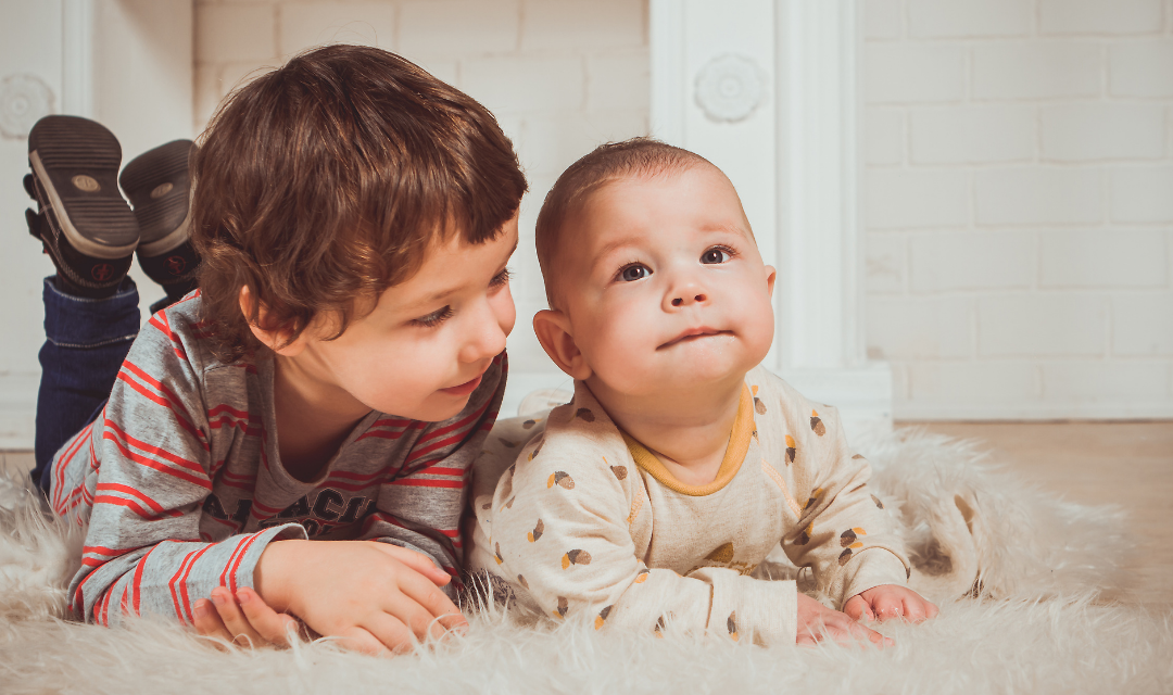 Young child smile at a baby as they play on a rug