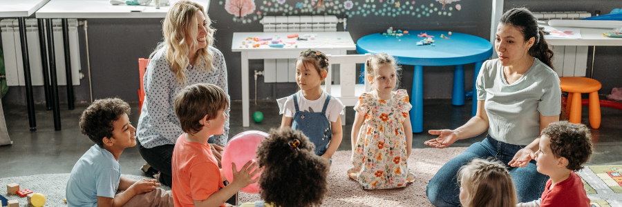 Two teachers sit with young children in a circle tossing a large pink ball