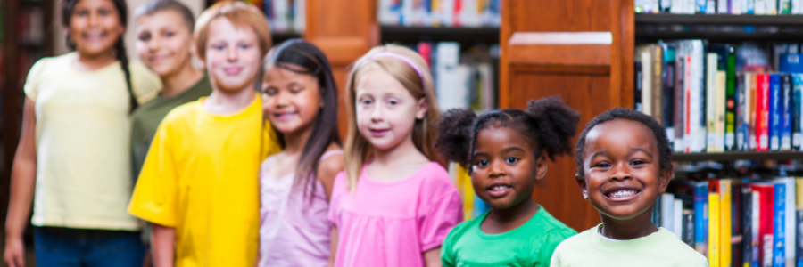 Children of various ages lined up in a library in front of a stack of books