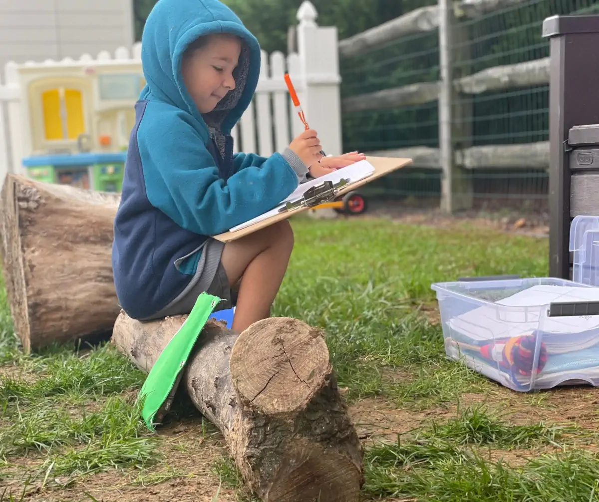 Child sitting on a log writing observations about a lesson in an outdoor classroom.
