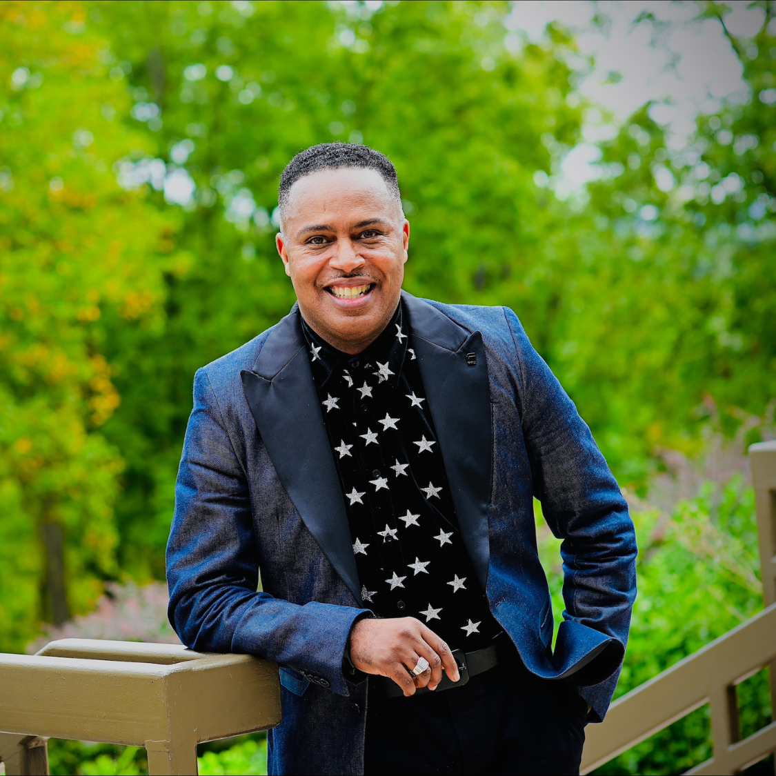 Dr. Moore stands smiling in a blue suit jacket and shirt with white stars with a background of trees behind him.