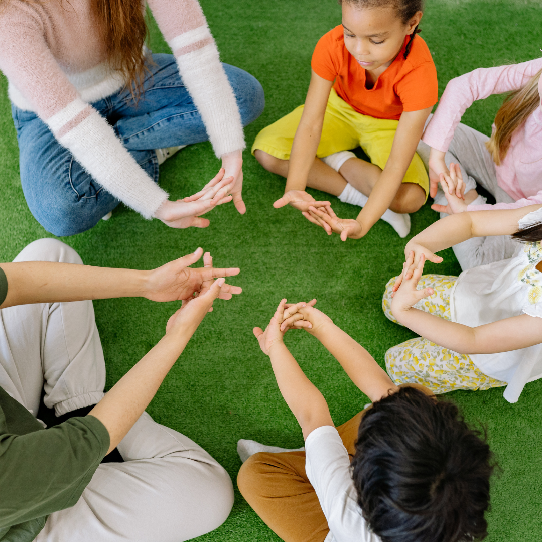 Overhead view of children and adults sitting in a circle, playing a hand-stretching game.
